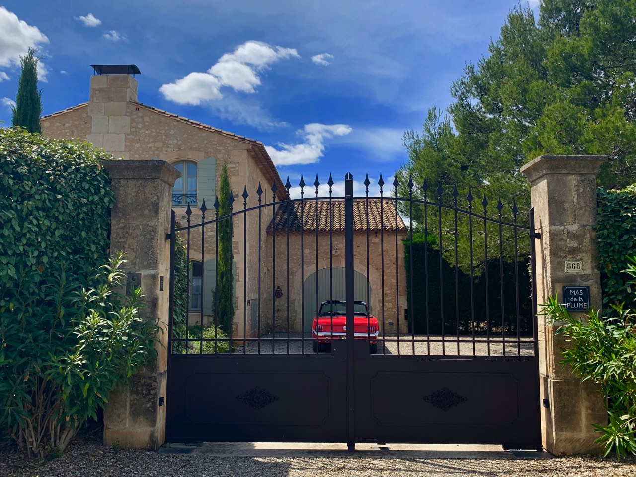 A red convertible parked behind ornate black gates in front of an elegant stone building surrounded by lush greenery under a clear blue sky.